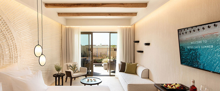 In der Suite des Mitsis Selection Rinela dominieren helle Sand- und Beigetöne, die den Raum warm und ruhig wirken lassen, mit Blick auf den Balkon.