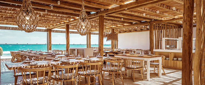 Auf der Holzterrasse des Outdoor-Restaurants öffnet sich der Blick über das ruhige Meer, während warme Naturtöne und offene Strukturen die Szenerie umrahmen.