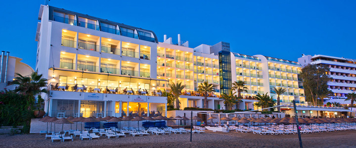 Außenansicht des Hotel Yalihan Una bei Nacht, vom Strand aus aufgenommen, das hell beleuchtete Gebäude leuchtet vor dem dunkelblauen Himmel.