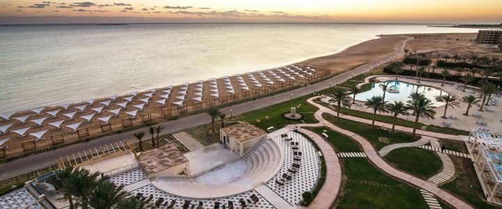 Vom Hotel eröffnet sich der Blick über die weitläufige Poollandschaft und den grünen Park bis hin zum Strand mit Sonnenliegen, dahinter liegt das Rote Meer im warmen Licht des Sonnenuntergangs.