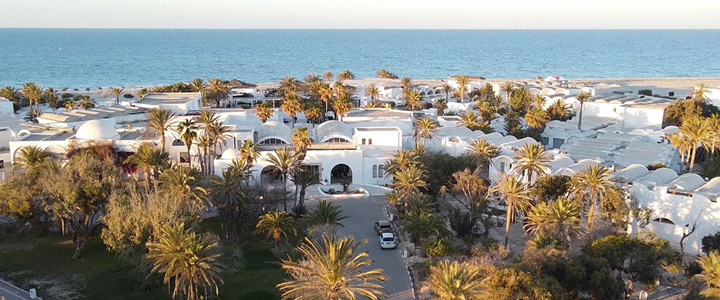 Sonne und Meer prägen den Blick über die Anlage mit weißen, mediterranen Häusern und Palmen, während sich das Mittelmeer ruhig am Horizont öffnet.